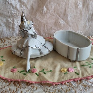 Puede incluir: Una caja de porcelana blanca con ribete plateado. La tapa presenta una figurita de una mujer con un vestido blanco y un sombrero puntiagudo. La caja est&aacute; sobre una tela bordada de color crema.