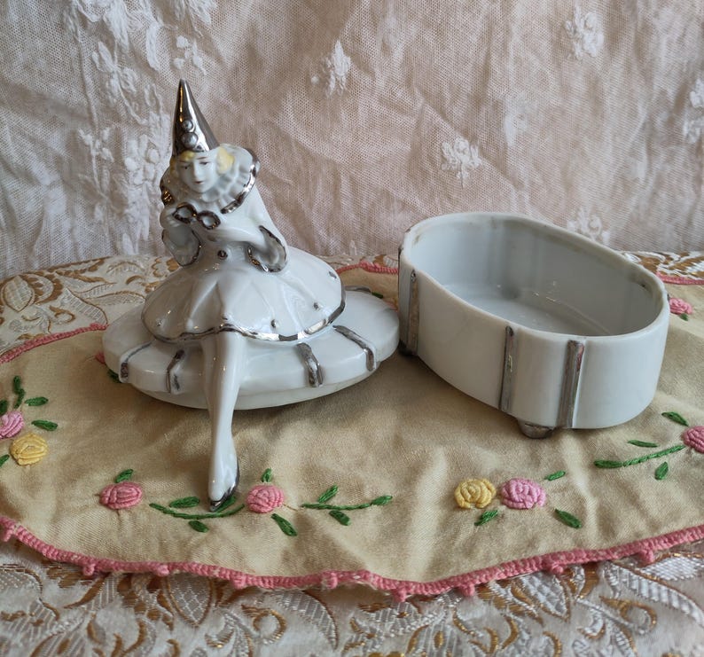 Puede incluir: Una caja de porcelana blanca con ribete plateado. La tapa presenta una figurita de una mujer con un vestido blanco y un sombrero puntiagudo. La caja est&aacute; sobre una tela bordada de color crema.
