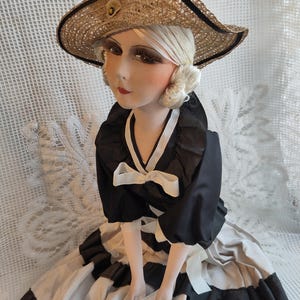 May include: A vintage doll wearing a straw hat, a black top with white bows, and a black and white dress. The doll has a painted face with red lips. The hat has a black trim and a small flower. The doll is in a seated position.
