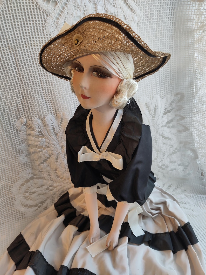 May include: A vintage doll wearing a straw hat, a black top with white bows, and a black and white dress. The doll has a painted face with red lips. The hat has a black trim and a small flower. The doll is in a seated position.