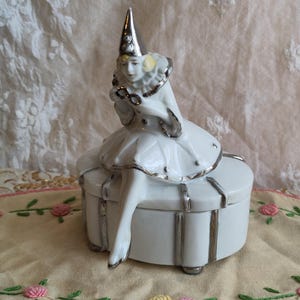 Puede incluir: Una caja de porcelana blanca con ribete plateado. Una figurita de porcelana de una mujer vestida de buf&oacute;n se sienta encima de la caja.
