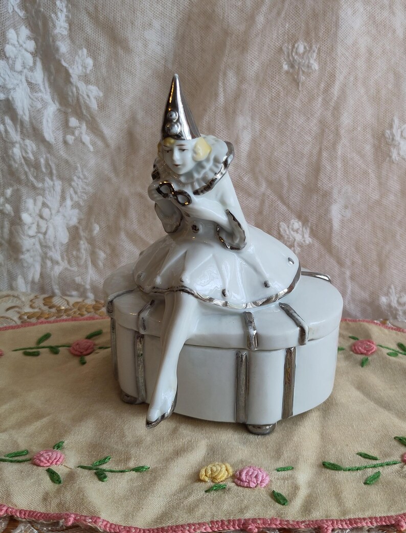 Puede incluir: Una caja de porcelana blanca con ribete plateado. Una figurita de porcelana de una mujer vestida de buf&oacute;n se sienta encima de la caja.