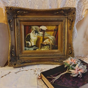 May include: A framed oil painting of three cats in a room. The cats are white, black, and gray. The painting is in a gold frame with ornate details.