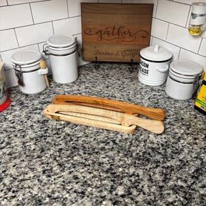 May include: Two wooden serving tongs, one light and one dark, rest on a speckled countertop. In the background are white ceramic canisters, a wooden cutting board with the word "gather" and the names "Dorina & Gary" engraved, and other kitchen items.