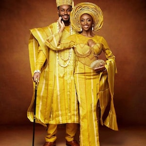 May include: A couple in traditional yellow Nigerian attire. The man is wearing a long-sleeved robe with vertical stripes and a matching cap. The woman is wearing a long-sleeved dress with vertical stripes and a matching head wrap. The man is holding a walking stick.