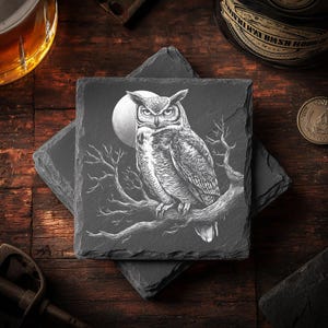 May include: Square slate coasters with an engraved owl design. The owl is perched on a tree branch in front of a full moon. The coasters are dark gray and rest on a wooden surface.