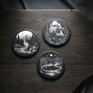 May include: Three round slate coasters with engraved wildlife designs. One features a rabbit in a forest, another a boar, and the third an otter in a pond. The coasters are black with white illustrations, set on a dark wooden surface.