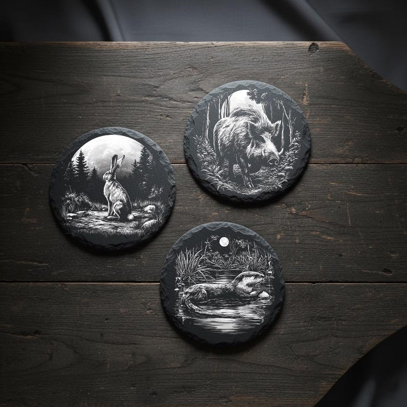 May include: Three round slate coasters with engraved wildlife designs. One features a rabbit in a forest, another a boar, and the third an otter in a pond. The coasters are black with white illustrations, set on a dark wooden surface.