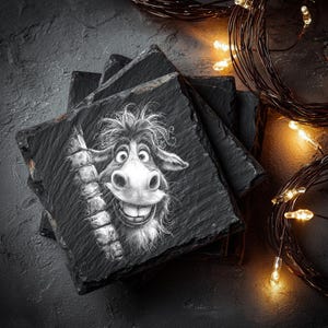 May include: A stack of square slate coasters with a whimsical, black and white illustration of a smiling donkey. The donkey's face is detailed with expressive eyes and a wide grin. The coasters are stacked on a dark surface, with warm fairy lights in the background.