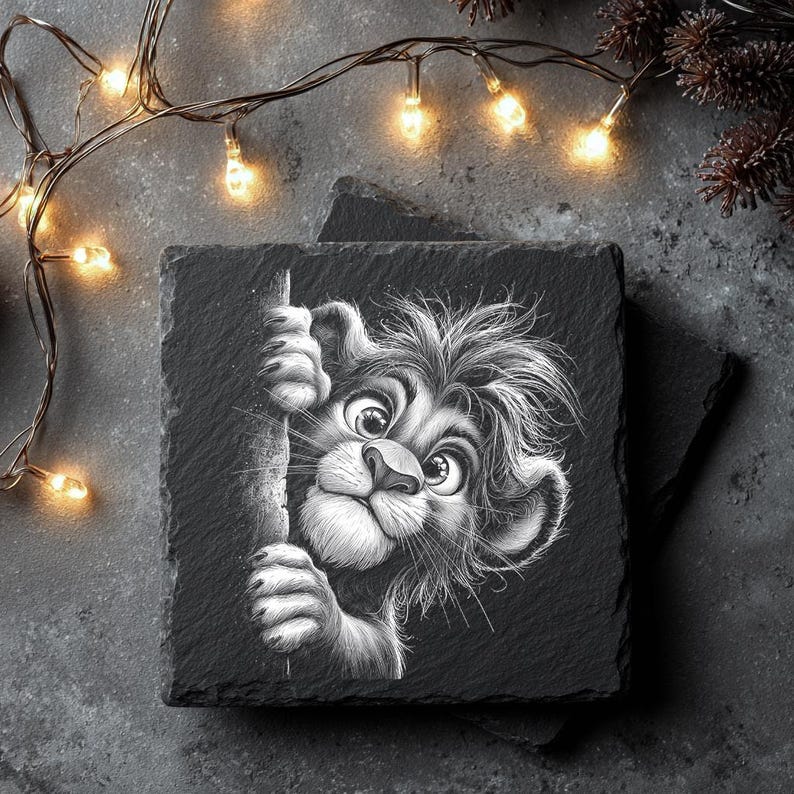 May include: Square black slate coaster featuring a detailed black and white illustration of a cartoon lion cub peeking around a tree. The lion has large eyes and a fluffy mane. The coaster is on a dark gray surface, with string lights and pine branches in the background.