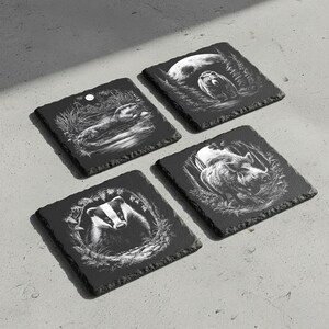 May include: Four square black slate coasters with etched wildlife designs. Each coaster features a different animal: an otter, a badger, a wild boar, and an owl. The designs are in white, set against the dark slate.