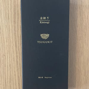 May include: A rectangular black box featuring gold text and a gold bowl icon. The text reads "金継ぎ Kintsugi", "TSUGUKIT", and "初心者 Beginner". The box has a smooth, matte finish and is oriented vertically, displaying branding for a kintsugi repair kit.