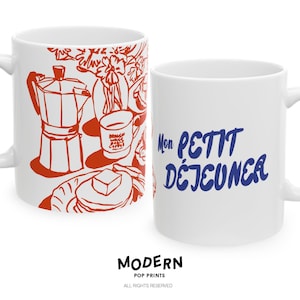 May include: Two white ceramic mugs. One mug features a red illustration of breakfast items, including a coffee maker, coffee cup, and pancakes. The other mug has the text "Mon PETIT DÉJEUNER" in blue. Both mugs have a handle.