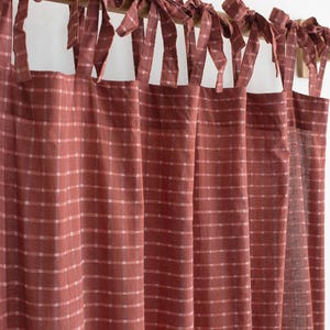May include: A terracotta-colored curtain featuring a white grid pattern. The curtain is suspended from a wooden rod by multiple fabric ties that are knotted at the top, creating a gathered, pleated effect across the length of the fabric panel.