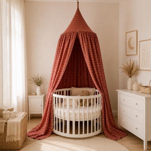 May include: A round white wooden cot positioned beneath a terracotta-coloured fabric canopy. The canopy features a subtle white grid pattern and hangs from a central point, draping down to the floor around the circular cot frame.