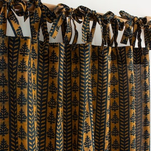 May include: A curtain panel featuring a repeating pattern of dark navy blue vertical stripes and stylised botanical motifs on a mustard yellow background. The curtain is suspended from a wooden rod by multiple fabric ties knotted at the top.