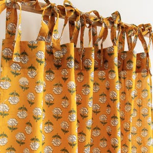 May include: A mustard yellow curtain panel featuring a repeating white floral pattern with green stems and leaves. The curtain is suspended from a wooden rod by multiple fabric ties knotted at the top, creating a gathered, pleated appearance.