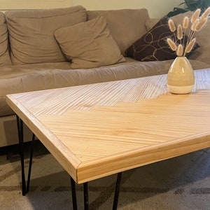 May include: A light wood coffee table with a chevron pattern top and black hairpin legs. The table is sitting on a beige rug.