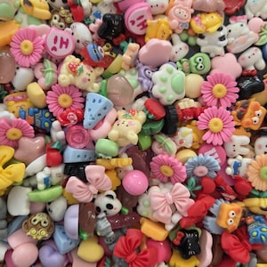 May include: A collection of colorful resin cabochons featuring various shapes including pink daisies, light blue cheese wedges, pastel bows, animal faces, and paw prints. The assortment includes small decorative charms in shades of pink, blue, yellow, and white.