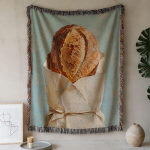 May include: A woven wall tapestry featuring a light blue background with a central illustration of a round, golden-brown loaf of crusty bread wrapped in a tan paper bag tied with string. The rectangular textile has a decorative fringed edge.