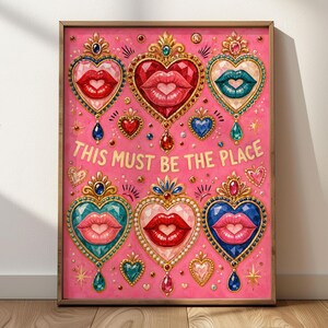 May include: A rectangular wall art print in a wooden frame featuring a vibrant pink background with six ornate heart-shaped motifs. Each heart contains a pair of lips and is decorated with jewels. The text THIS MUST BE THE PLACE is centered.