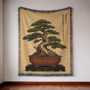 May include: A woven throw blanket featuring a detailed illustration of a green bonsai tree in a red decorative pot on a black stand. The background is a tan color with the text "Japanese characters" printed vertically in black on the right side.