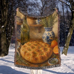 May include: A woven throw blanket featuring a still life print of a round loaf of focaccia bread, a glass bottle of olive oil, a blue and white patterned ceramic vase with rosemary sprigs, two oranges, and a small bowl of herbs on a wooden table.