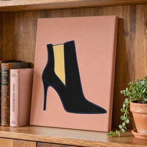 May include: A rectangular canvas print featuring a stylized illustration of a black high-heeled ankle boot with a yellow elastic side panel. The boot is centered on a solid, muted terracotta-colored background, creating a clean and graphic visual.