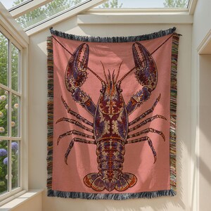 May include: A woven throw blanket featuring a large, detailed lobster graphic in shades of purple, orange, and dark blue against a solid dusty pink background. The blanket has a decorative fringe border with multicolored threads on all four sides.
