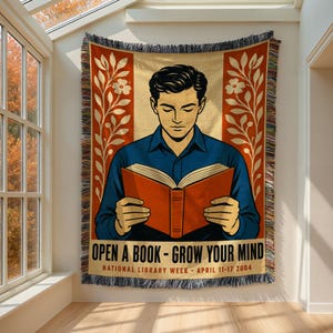 May include: A woven throw blanket featuring a graphic illustration of a person reading an orange book. The design includes floral borders, the text "OPEN A BOOK - GROW YOUR MIND" and "NATIONAL LIBRARY WEEK - APRIL 11-17 2004" with fringed edges.