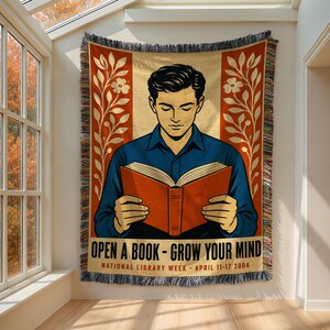May include: A woven throw blanket featuring a graphic illustration of a person reading an orange book. The design includes floral borders, the text "OPEN A BOOK - GROW YOUR MIND" and "NATIONAL LIBRARY WEEK - APRIL 11-17 2004" with fringed edges.