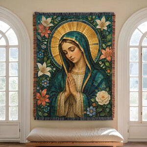May include: A woven tapestry wall hanging featuring a portrait of a figure with hands clasped in prayer, wearing a teal head covering and a gold halo. The design is surrounded by white and orange lilies and small blue flowers with a fringed edge.