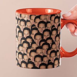 Peut inclure: Une tasse à café en céramique avec une anse et un intérieur rouges. L'extérieur de la tasse est recouvert d'un motif répété d'un visage. La tasse est tenue par une main.