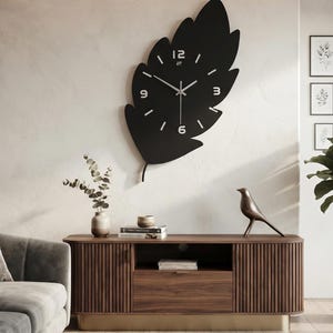 Pode incluir: Um grande relógio de parede preto fosco em forma de folha, com marcadores de hora e números brancos nas posições de 12, 3, 6 e 9 horas. O relógio possui ponteiros finos de cor prateada para horas, minutos e segundos no centro.