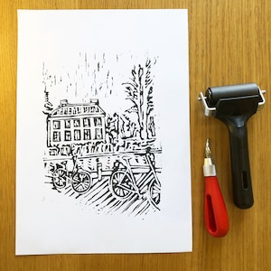 May include: Black and white linocut print of a building with windows, trees, and bicycles. The print is on white paper and is framed by a white mat.