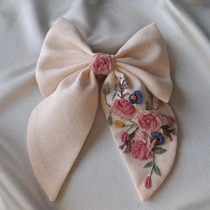 Peut inclure: Un grand nœud en tissu rose pâle avec des détails floraux brodés. La broderie présente des fleurs roses, bleues et jaunes avec des feuilles vertes. Accessoire pour cheveux.