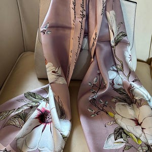 May include: A mauve silk scarf with a floral pattern of large white flowers with pink and red centers. The scarf has a border with the alphabet printed on it. The scarf is draped over a white handbag.