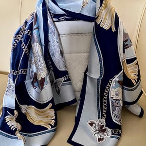 May include: A navy blue and light blue silk scarf with a floral and chain design. The scarf features a repeating pattern of flowers, tassels, and chains. The scarf is draped over a white handbag.