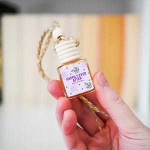 May include: A small, clear glass air freshener bottle with a wooden cap and a decorative string. The label reads "Happily Ever After" with floral and star designs. The bottle is held in a hand.