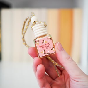 May include: A small, clear glass bottle air freshener with a wooden cap and a woven gold and beige string. The label reads "Main Street Parlor" with ice cream cone illustrations on a pink background. The bottle is held in a hand.