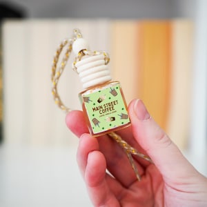 May include: A small, clear glass bottle with a light brown wooden cap and a decorative string. The label reads "Main Street Coffee" with coffee cup illustrations. The bottle is held in a hand.