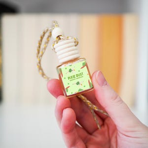 Op de afbeelding: Een kleine, heldere glazen fles luchtverfrisser met een houten dop en een goudkleurig touwtje. Het etiket leest "PIXIE DUST" met een groene achtergrond en illustraties. De fles wordt in een hand gehouden.