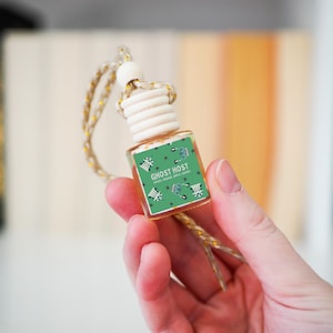 May include: A small, clear glass bottle with a wooden cap and a gold-coloured string for hanging. The label reads "GHOST HOST" with a green background and decorative elements. The bottle is held in a hand.
