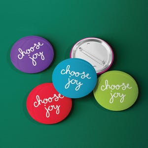 May include: Four round buttons with the phrase "choose joy" written in white cursive on a colored background. The buttons are purple, blue, red, and green.