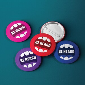 May include: Four round buttons with the text "BE HEARD" in white letters on a colorful background. The buttons are pink, red, purple, and blue. Each button features a stylized mouth design.
