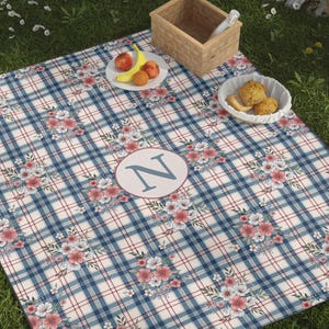 May include: A picnic blanket with a blue and white plaid pattern, accented with floral designs and a circular monogram with the letter "N". The blanket is on a grassy surface, with a soccer ball, tennis racket, and food items.