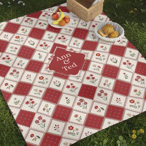Peut inclure: Une couverture de pique-nique à carreaux rouges et blancs avec des motifs floraux et le texte "Ann & Ted" sur un carré rouge. La couverture est sur l'herbe, avec un ballon de foot, une raquette de tennis et un panier de pique-nique.