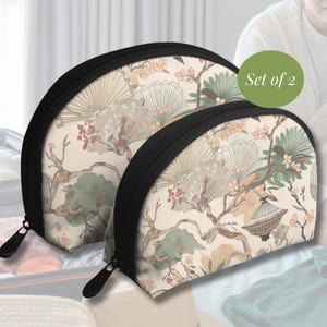 Peut inclure: Un ensemble de deux trousses de toilette semi-circulaires avec un fond crème et un motif répétitif d'éventails, de branches d'arbres et de petites fleurs roses. Chaque trousse possède une fermeture éclair noire et un bord noir.