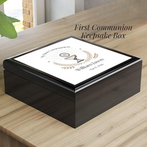May include: A black keepsake box with a white top featuring the text "My First Communion" and "William Jason" with a date. The box has a glossy finish and a decorative design of a chalice and wheat. The words "First Communion Keepsake Box" are also on the top.