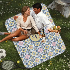 May include: A picnic blanket with a blue, yellow, and pink floral pattern. The blanket is on a grassy surface with a soccer ball and tennis racket nearby. A couple is sitting on the blanket, enjoying a picnic with wine and fruit.